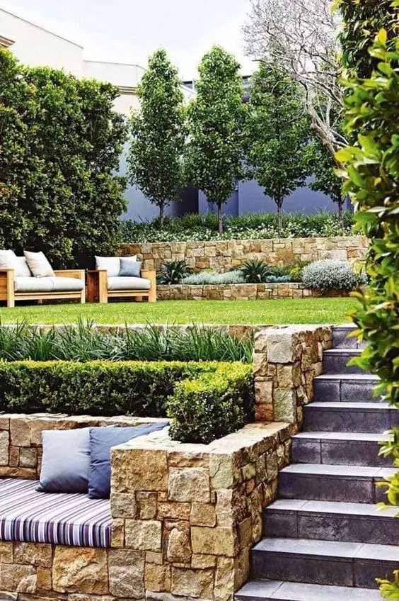 a two level outdoor space with a sitting zone in the lower part some greenery and a green lawn with outdoor furniture and some plants on top