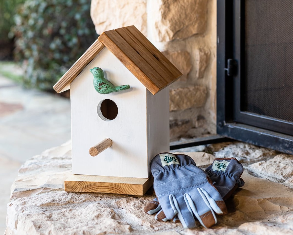 The Dreamy Farmhouse Cottage white birdhouse with a dark roof and a small perch.