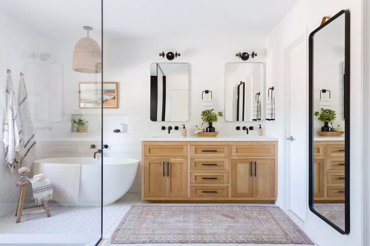 Bright bathroom with a light oak double vanity, twin rectangular mirrors, black sconces, a soaking tub behind glass, and a Persian-style rug.
