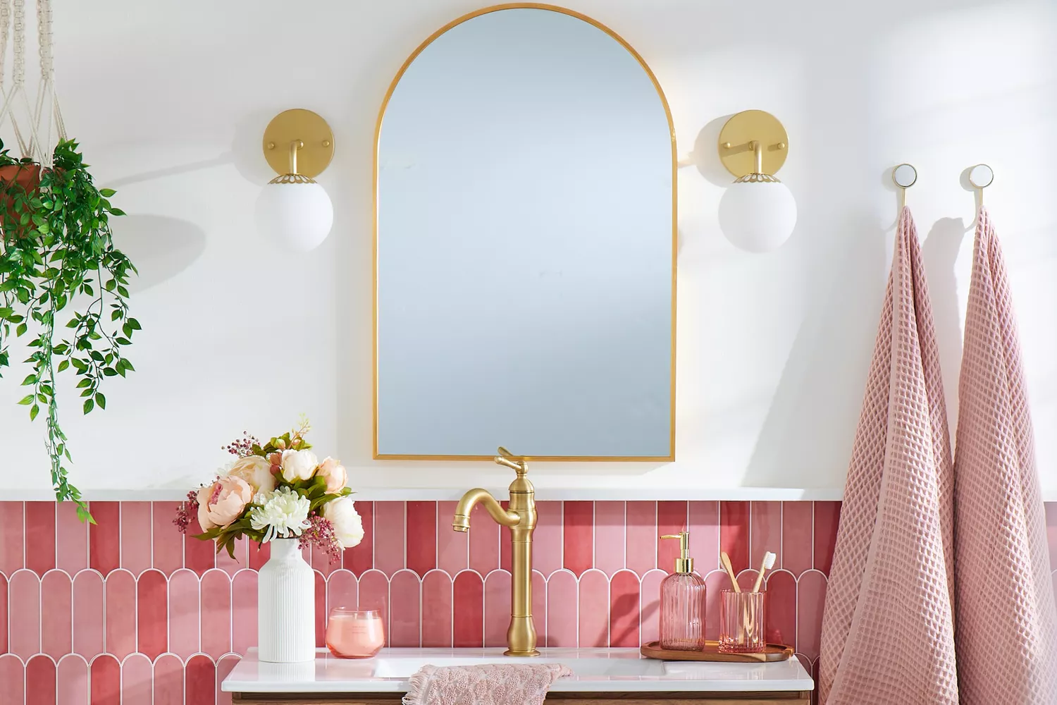 Vanity area with pink and rose scalloped tile backsplash, arched gold mirror, globe sconces, and vintage-style brass faucet.