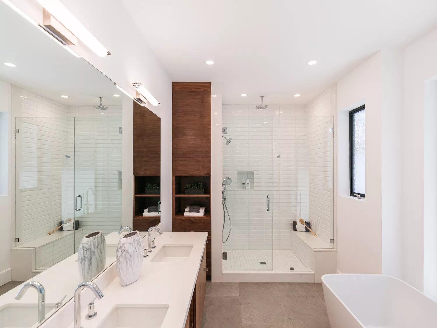 Large modern bathroom with a white brick-tile walk-in shower, seamless glass door, long double vanity, and tall wood storage cabinet.