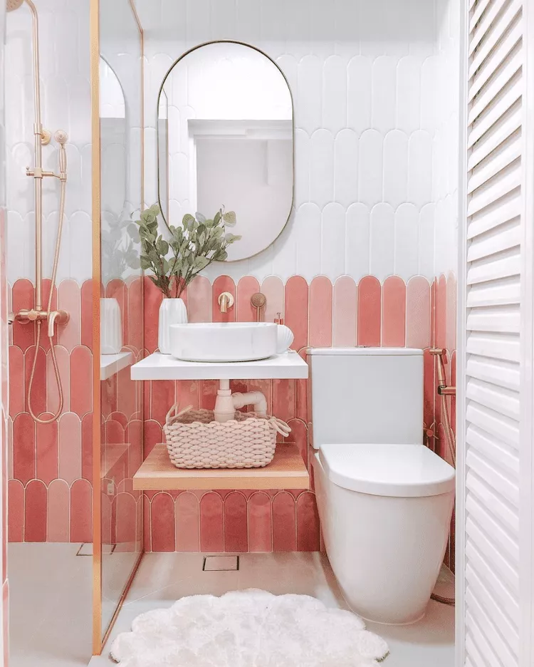 Compact bathroom with vertical pink scalloped tiles, floating white sink, wood shelf, rose gold fixtures, and a round mirror.