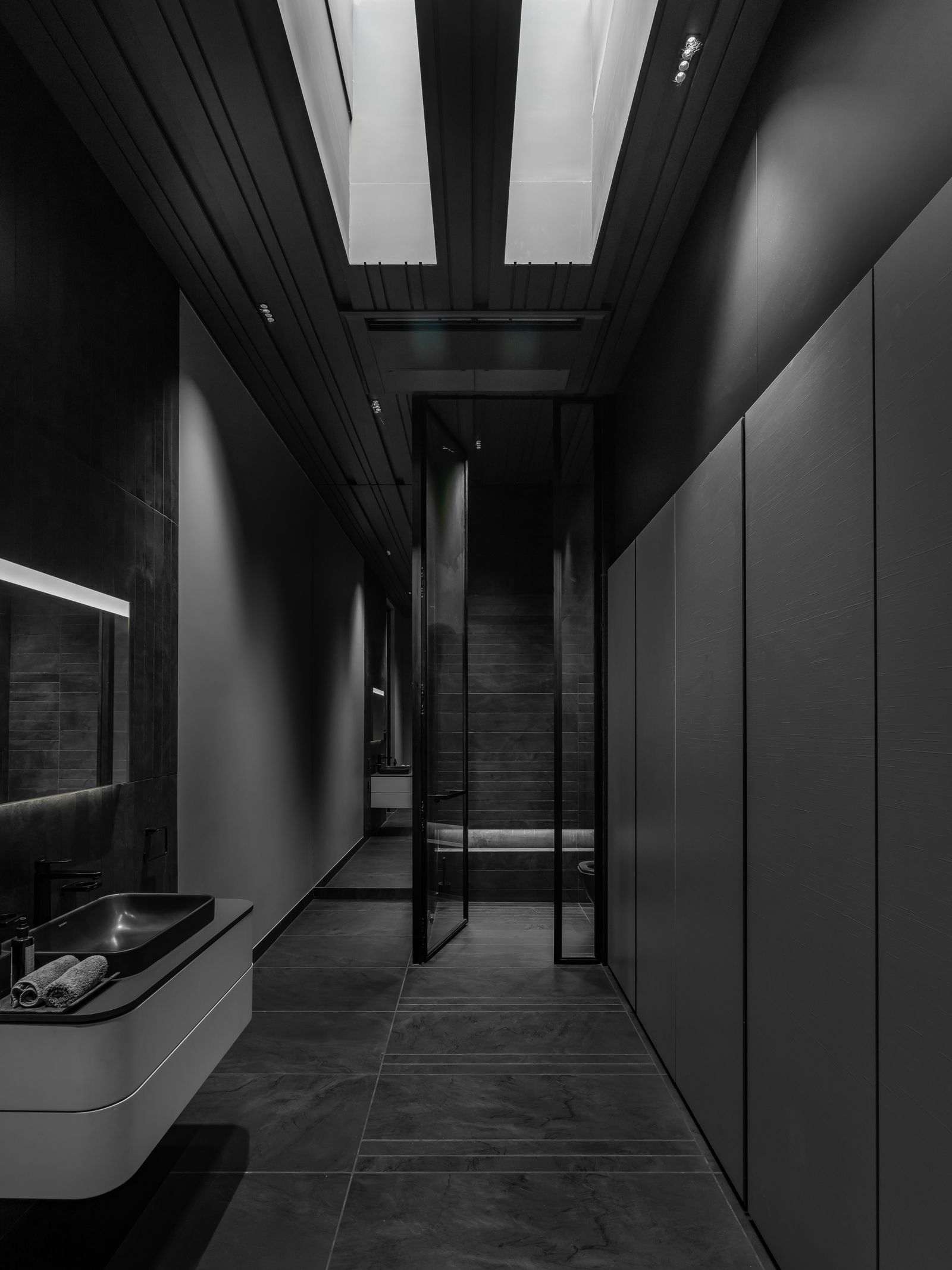 Modern small bathroom with dark large-format floor tiles, sleek handle-less cabinetry, wall-mounted vanity, and a skylight creating dramatic natural light.