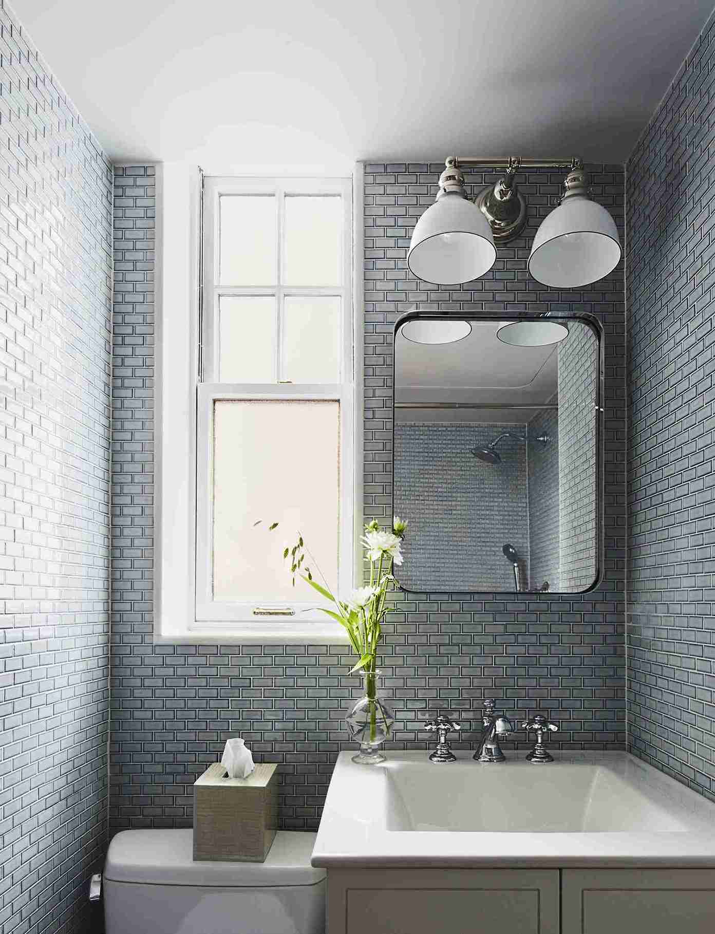 Small bathroom with slate-blue subway tiles covering walls, white pedestal sink, bright window, chrome faucet, and classic white wall sconces.
