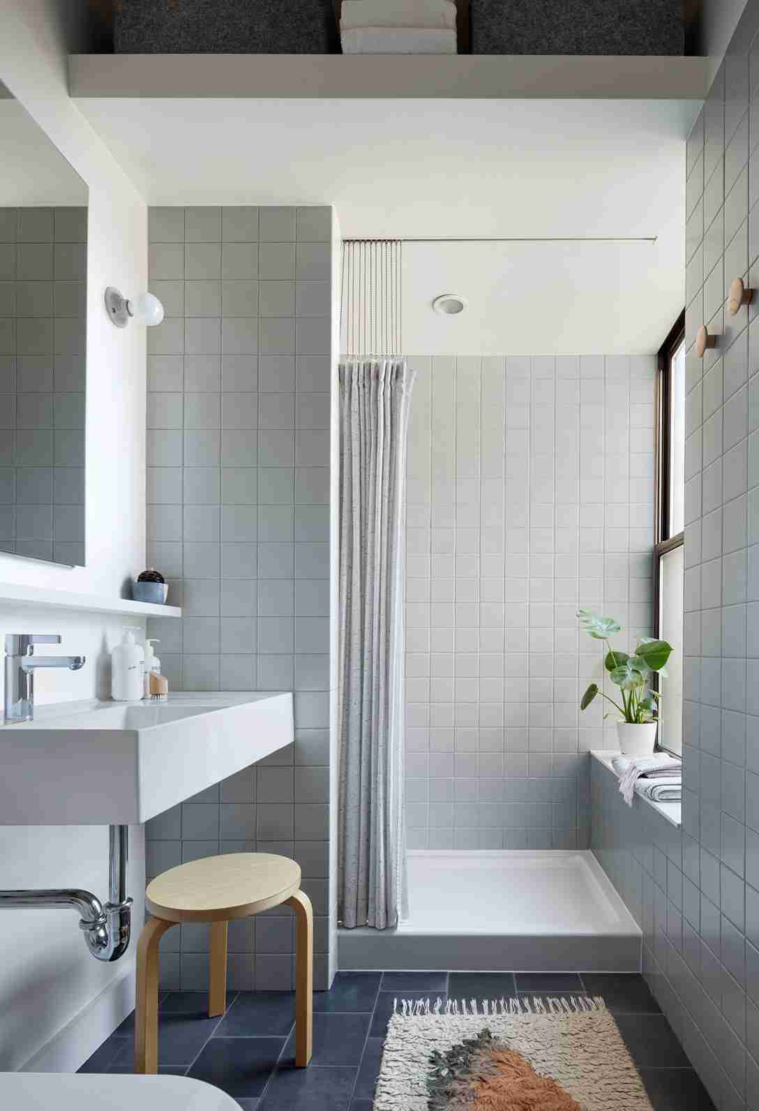 Small bathroom with large square gray wall tiles, floating white trough sink, wooden stool, high shelf with baskets, and a textured rug on dark flooring.