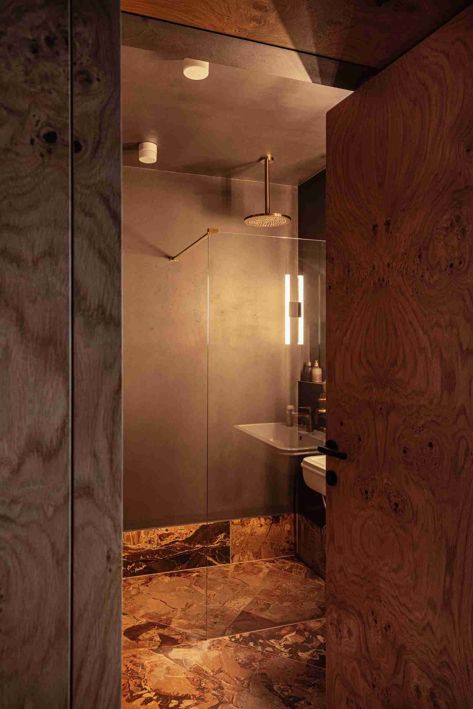 Small bathroom with warm brown veined marble flooring extending into shower, wood-grain door and ceiling, and soft lighting from wall sconce and recessed fixtures.