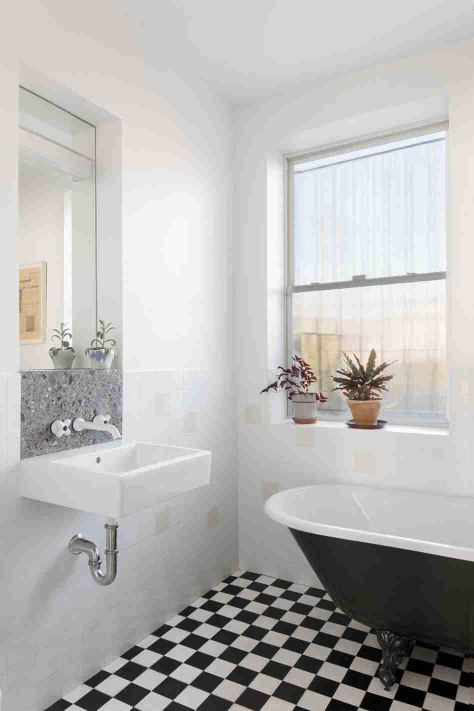 Small bathroom with large-scale terrazzo tiles on walls and floor, wood-paneled ceiling, matte black freestanding tub, black floor-mounted tub filler, and wooden stool.